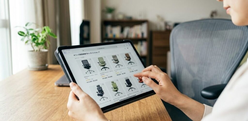 自宅にて、タブレット端末でオフィス家具専門のオンラインショップのウェブサイトを見ている日本人女性の手元。様々なオフィスチェアが並んでいる。

