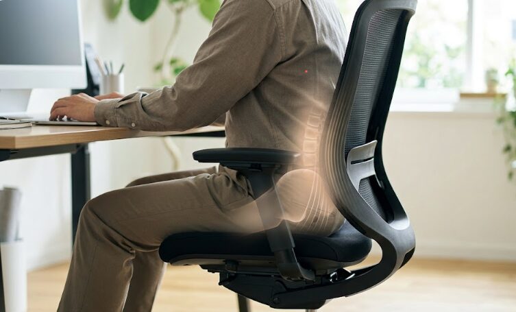 コクヨingチェアの360°グライディング座面が、デスクワーク中の日本人男性の骨盤を優しくサポートし、腰痛への負担を軽減する様子を斜め後方から捉えたクローズアップ。動く座面と背もたれの連携が視覚化されている。